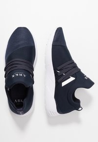 Mörkblå sportskor med en slät yta, vävda mesh-accenter, framträdande snörningssystem och en vit gummisula. Varumärket visas.