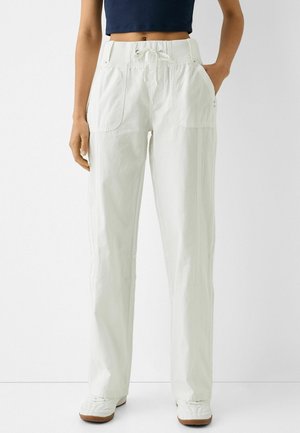 Femme portant un pantalon blanc à jambes larges avec une taille élastique à cordon, des poches plaquées sur le devant, associé à des baskets blanches et un top court bleu marine.