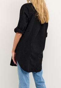 Chemise noire à manches longues avec boutons, manches retroussées, ourlet légèrement incurvé et coupe décontractée, associée à un jean bleu clair.
