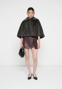 Dunkelgrauer Kunstfellumhang mit Kragen, getragen über einem burgunderfarbenen Leder-Minirock. Eine schwarze Handtasche und spitze schwarze Schuhe vervollständigen das Outfit.