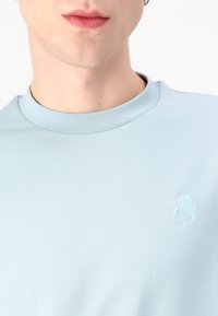 Γαλάζιο μπλουζάκι με λαιμόκοψη φτιαγμένο από μαλακό ύφασμα, με μικρό κεντημένο σχέδιο στο στήθος, ομαλή υφή και απλή σιλουέτα.