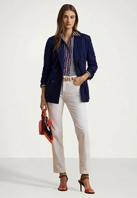 Γυναίκα με μπλε ριγέ σακάκι, ριγέ πουκάμισο, λευκό παντελόνι, πορτοκαλί τσάντα με φουλάρι και πορτοκαλί ψηλοτάκουνα πέδιλα, να στέκεται μπροστά σε λευκό φόντο.