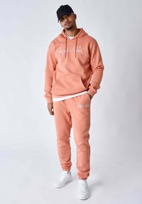 Sudadera con capucha rosa y pantalones deportivos a juego hechos de tejido suave, con texto de logo en blanco. Combinados con zapatillas blancas.