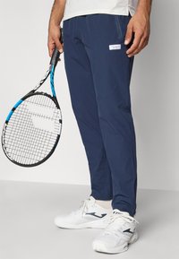 Ναυτικές αθλητικές παντελόνες με τσέπη που κλείνει με φερμουάρ, σε συνδυασμό με λευκά και ναυτικά αθλητικά παπούτσια. Μια ρακέτα του τένις κρατιέται στο αριστερό χέρι.