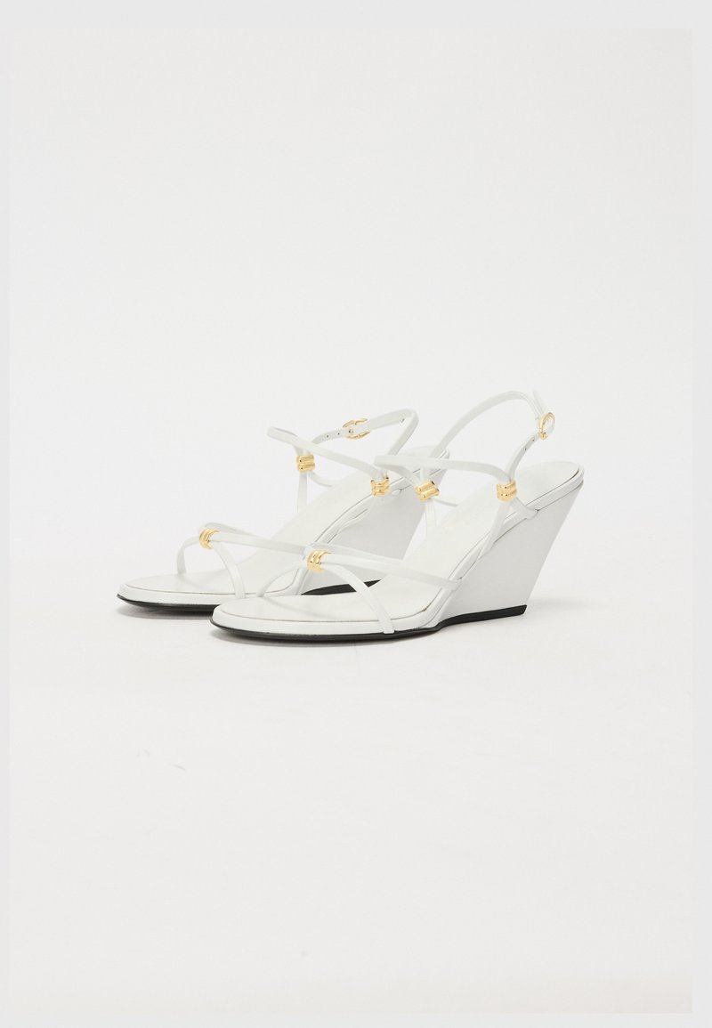White wedge sandals with thin straps, gold accents, a smooth finish, and a black sole; minimalist design with a tapered wedge shape.