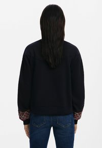 Felpa nera con maniche ricamate che presentano un motivo geometrico. Indossata con jeans blu in denim, vista da dietro. Tessuto liscio.