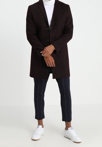 Abrigo de lana burdeos con dos botones, mangas largas y bolsillos laterales, combinado con pantalones azul marino a rayas y zapatillas blancas.