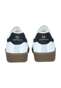 Fehér sportcipők fekete részletekkel, sima bőr felsőrésszel, kerek orral és barna gumitalppal. A logó jól láthatóan a sarka mögött helyezkedik el.