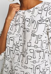 Blusa bianca con stampa di un volto astratto nero; presenta un collo rotondo e maniche corte; texture liscia.