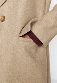 Abrigo de lana beige con cierre de botones, bolsillo lateral y puño de punto burdeos asomando por la manga. Textura suave y diseño ajustado.