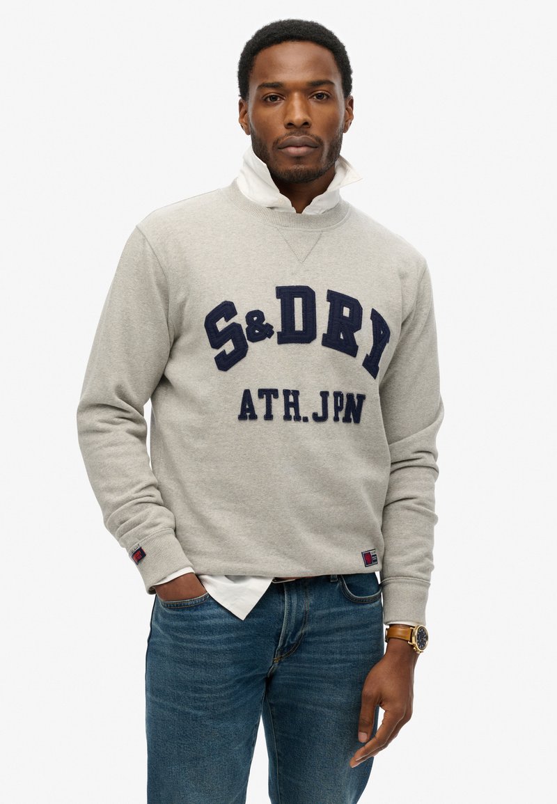 Sudadera gris con letras en bloque azul marino "S&D" y "ATH.JPN" en la parte delantera. Combinada con unos jeans de color azul claro y una camisa blanca con cuello.