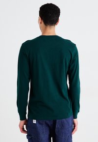Langarm, donkergroen shirt van zachte stof. Ronde halslijn, gladde textuur en eenvoudig ontwerp. Geen zichtbare patronen of bevestigingen.
