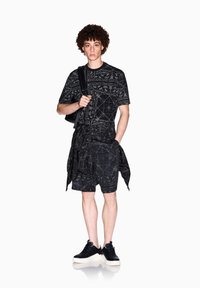 Maglietta grafica nera con motivi intricati, abbinata a pantaloni corti neri e un dettagli in tessuto stratificato, completata da sneakers nere e uno zaino.