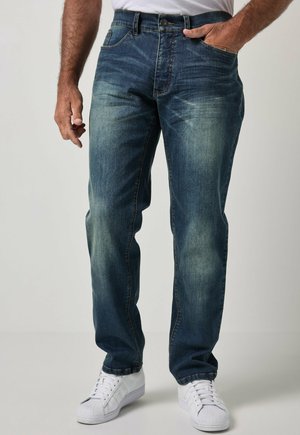 Hombre con vaqueros azules desgastados y zapatillas blancas, con una mano en el bolsillo y la otra relajada junto al cuerpo.