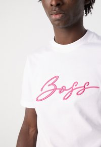 Valge puuvillane T-särk, millel on silmatorkav roosa broderdamisega "Boss" logo. Ümara kaeluse ja lühikeste varrukatega, sile tekstuur.
