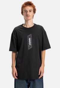 Černé oversize tričko vyrobené z bavlny, s bílým logo designem uprostřed. Tkanina vypadá hladce a má krátké rukávy.