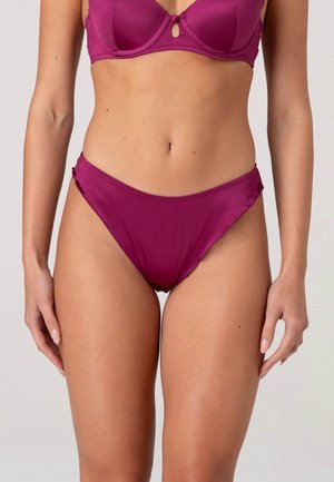 Donna che indossa un reggiseno magenta coordinato e slip bikini a vita alta, in piedi contro uno sfondo chiaro e uniforme, mani rilassate ai lati.