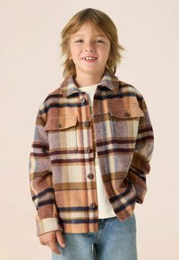 Flanellen overhemd met een geruit patroon in oranje, bruin en crème, met twee borstzakken en bruine knopen. Gedragen over een licht shirt.