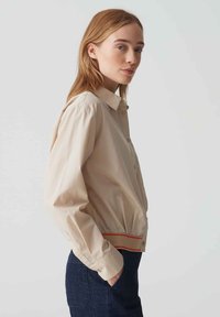 Beige overhemd met knopen en een kraag, elastische onderkant, en een rode accent aan de onderkant, gemaakt van een gladde stof. Casual pasvorm, lange mouwen.