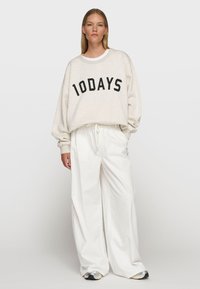 Hellbeige Sweatshirt mit "10DAYS" in fetten schwarzen Buchstaben, kombiniert mit lockeren weißen Hosen und grauen Sneakers. Weicher Stoff, lässiger Schnitt.