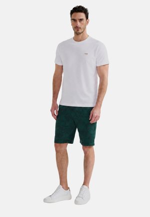 Uomo che indossa una maglietta bianca a maniche corte, pantaloncini verdi scuri con fantasia e sneakers bianche, in piedi su uno sfondo semplice.