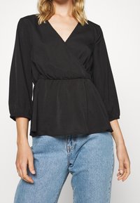 Blusa nera avvolgente con scollo a V, vita arricciata e maniche 3/4, abbinata a jeans a vita alta blu chiaro, tessuto dalla texture liscia.