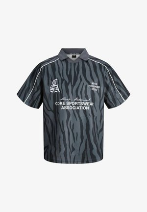 Garçon debout, portant un polo rayé zèbre noir et gris avec texte, pantalon large sombre, et baskets blanches, regardant sur le côté.