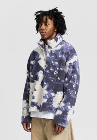 Fleece pullover in cream and navy tie-dye pattern, featuring a quarter-zip design and stand collar. Soft texture with relaxed fit.