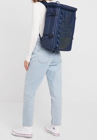Personne portant un jean bleu clair, un haut blanc, des baskets blanches montantes et un sac à dos rectangulaire bleu marine avec le logo "The North Face".