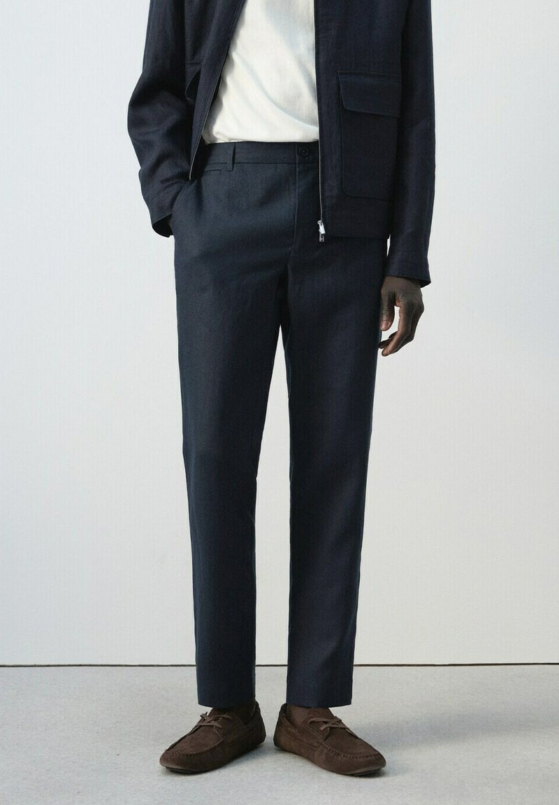 Uomo con pantaloni blu navy, scarpe scamosciate marroni, camicia bianca e giacca blu navy, in piedi contro uno sfondo chiaro e semplice.