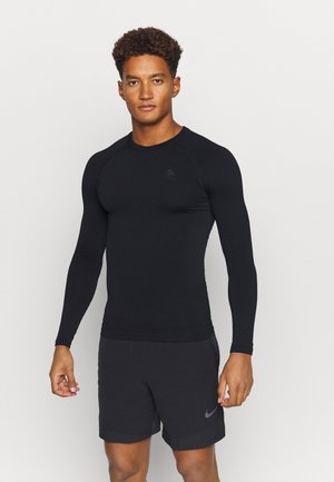 Schwarzes langärmliges Sportoberteil aus strukturiertem Material, figurbetontes Design und ein kleines Logo auf der Brust, kombiniert mit dunklen Shorts.