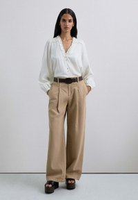Blusa blanca de botonadura completa con cuello con volantes, combinada con pantalones de talle alto beige con plisados. Sandalias de plataforma negras completan el look.
