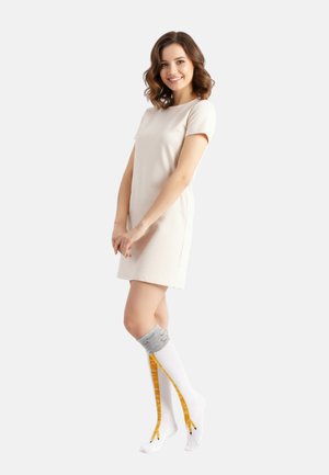 Junge Frau in einem kurzen beigen Kleid lächelt und trägt weiße Kniestrümpfe mit gelben Giraffenmuster und grauen Bündchen.