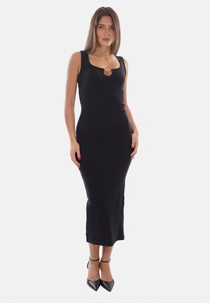 Femme portant une robe midi noire sans manches avec un détail anneau doré au niveau de l’encolure et des talons noirs à bout pointu et bride à la cheville.