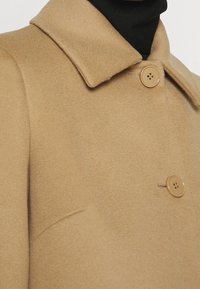 Manteau en laine couleur sable avec un col classique, doté de deux boutons ronds, une texture lisse et un détail de couture sur le côté.