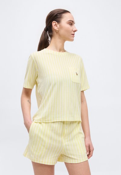 Γυναίκα με καστανή κοτσίδα, που φοράει κίτρινο και λευκό πουκάμισο με κάθετες ρίγες και κοντά μανίκια, καθώς και σορτς που ταιριάζουν, στέκεται με τα χέρια στις τσέπες.