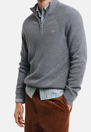 Man wearing gray quarter-zip sweater over light green striped shirt with brown corduroy pants, one hand in pocket, against plain background.