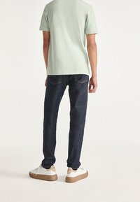 Ljusgrön kortärmad skjorta, mörkblå jeans och vita sneakers med bruna sulor; synliga sömmar och fickdetaljer på jeansen.