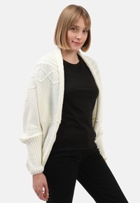 Junge Frau mit blonden Bob-Haarschnitt, die eine weiße Strickjacke über einem schwarzen Shirt und schwarzen Hosen trägt, vor einem schlichten hellen Hintergrund.