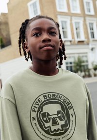 Šviesiai žalia tinklinė džemperis su juodu apskritu grafiniu dizainu, kuriame vaizduojamas kaukolės ir štangos motyvas bei užrašu „FIVE BOROUGHS“.