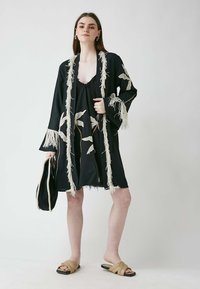 Svart kimono-stil klänning med fransdetaljer och vit blommig brodyr; lagerdesign; matchad med bruna slide-sandaler. Svart väska hålls.