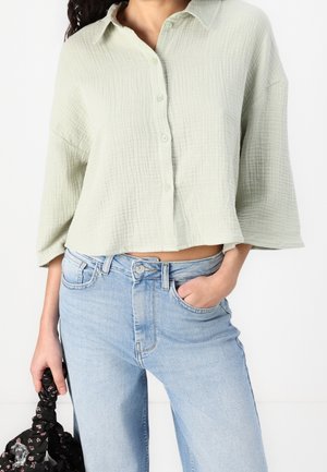 Kobieta w jasnooliwkowej, teksturowanej koszuli z rozkloszowanymi rękawami i jasnoniebieskich, wysokowasowych dżinsach, trzymająca czarną torbę w wzory.