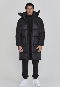 Schwarze Daunenjacke mit langem Design, ausgestattet mit großen Taschen, einem hohen Kragen und einem Reißverschluss vorne. Getragen über schwarzer Kleidung und weißen Schuhen.
