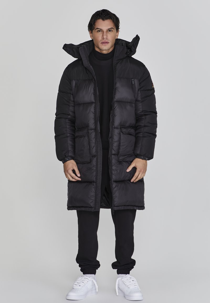 Schwarze Daunenjacke mit langem Design, ausgestattet mit großen Taschen, einem hohen Kragen und einem Reißverschluss vorne. Getragen über schwarzer Kleidung und weißen Schuhen.