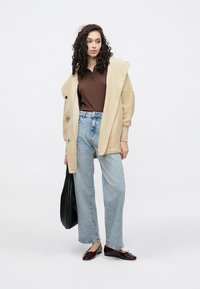 Casaco de fleece bege, polo castanha com riscas, jeans azul claro de perna larga e sapatos bordeaux. Modelo a segurar uma bolsa preta, fundo minimalista.