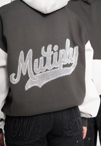Hanorac gri cu glugă și mâneci albe, având un logo „Multiply” argintiu brodat și pufos pe spate. Jeans negri cu litere brodate.