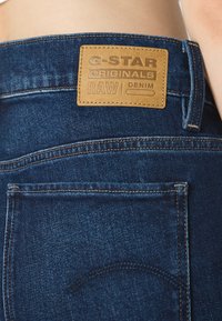 Temno modri jeans iz denima, ki imajo teksturirano usnjeno nalepko z reliefnim logotipom. Šivi so kontrastni in dobro opredeljeni.