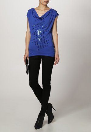 Vrouw draagt een blauw mouwloos topje met glanzende diagonale strepen, zwarte strakke broek, zwarte enkellaarsjes met hak, houdt een zwarte clutch vast.