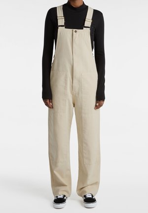 Beżowe bawełniane ogrodniczki z regulowanymi szelkami, przednimi kieszeniami, szerokimi nogawkami i zapinane na guziki. Noszone z czarną koszulką z długim rękawem i trampkami.