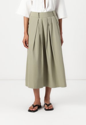 Vrouw die een beige geplooide midi-rok draagt, een wit blouse met wijde mouwen en zwarte platte sandalen, staande op een witte achtergrond.